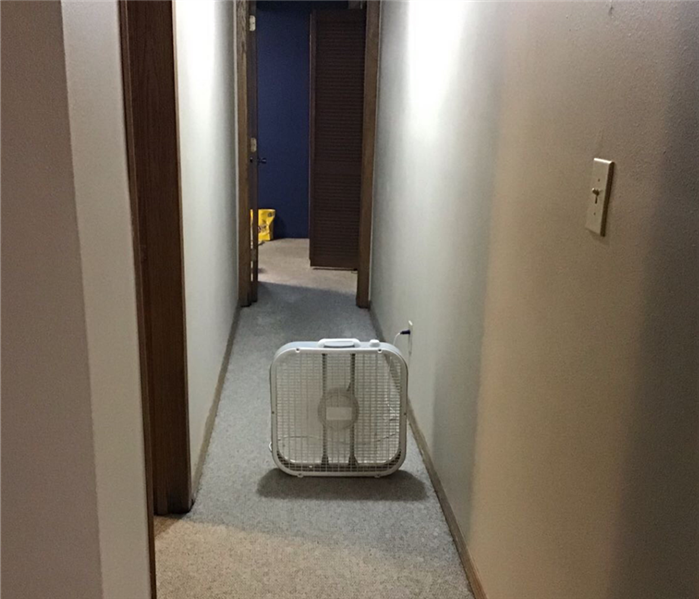 A hallway with white walls on each side and one white box fan in the middle of the hallway.